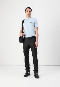 Light blue short-sleeve t-shirt with a small graphic, paired with black textured trousers and black shoes. Black shoulder bag is worn crossbody.