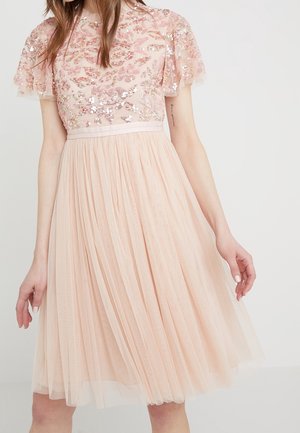 Femme portant une robe rose clair mi-longue, avec un haut floral à sequins et des manches courtes transparentes, debout devant un fond blanc uni.