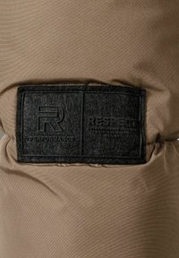 Una tela marrón con una textura suave presenta una etiqueta de fieltro negra que muestra "R PERFORMANCE" y "RESPECT" en letras en relieve.