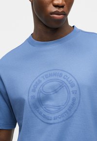 Άνδρας που φοράει γαλάζιο μπλουζάκι με ανάγλυφο κυκλικό λογότυπο που απεικονίζει μια μπάλα του τένις και το κείμενο "BOSS TENNIS CLUB 97 ESTABLISHED SPORTS BRAND."