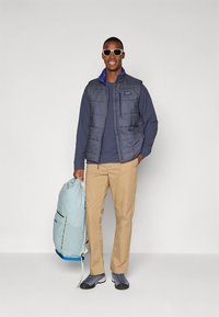 Siva puhasta jakna brez rokavov čez modro majico z dolgimi rokavi, bež hlače in sivi čevlji; nosi svetlo modro nahrbtnik; nosi sončna očala.