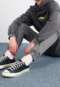 Donkergrijze hoodie met logo, lichtgrijze elastische broek en donkergroene canvas sneakers met witte veters en rubberen zolen. Roze sokken.