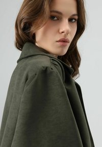 Veste vert olive en tissu doux et texturé. Comprend un col et des manches bouffantes, montrant un gros plan du design.