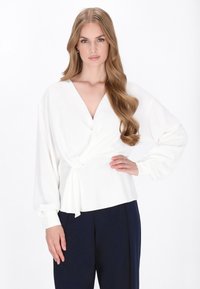Blusa bianca avvolgente con scollo a V profondo, maniche lunghe a sbuffo e dettaglio annodato in vita. Tessuto liscio con dettagli a bottoni sui polsini.