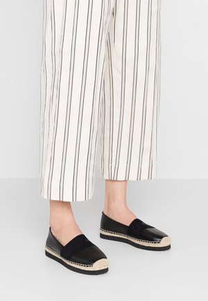 Jambes inférieures portant un pantalon blanc à rayures noires verticales et des chaussures noires sans lacets avec une bordure tressée beige sur les semelles.