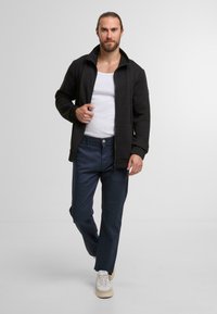 Mann geht vorwärts, trägt ein weißes Tanktop, eine schwarze Zip-Jacke, dunkelblaue Hosen und weiße Sneakers vor einem einfarbigen grauen Hintergrund.