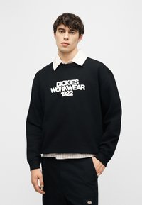 Jeune homme portant un sweat-shirt noir avec le texte « Dickies Workwear 1922 », une chemise blanche à col en dessous, les mains dans les poches de son pantalon noir.