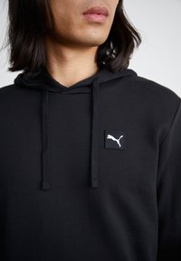 Persona che indossa un felpa nera con cappuccio e lacci e un piccolo logo Puma bianco sulla parte superiore sinistra del petto, su uno sfondo semplice.