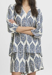 Ensemble blouse ample et short en crème, présentant un motif damassé bleu complexe. La blouse possède un col en V et des manches trois-quarts.