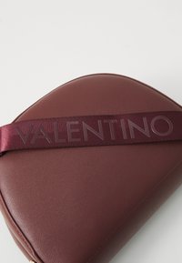 Bordeaux lederen handtas met een gladde textuur, voorzien van een brede satijnen schouderriem met de label "VALENTINO" in tonale letters. Ronde vorm.