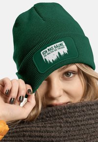 Green knit beanie with a textured finish. Features a rectangular patch with white lettering and forest graphic. Worn just above the brow.