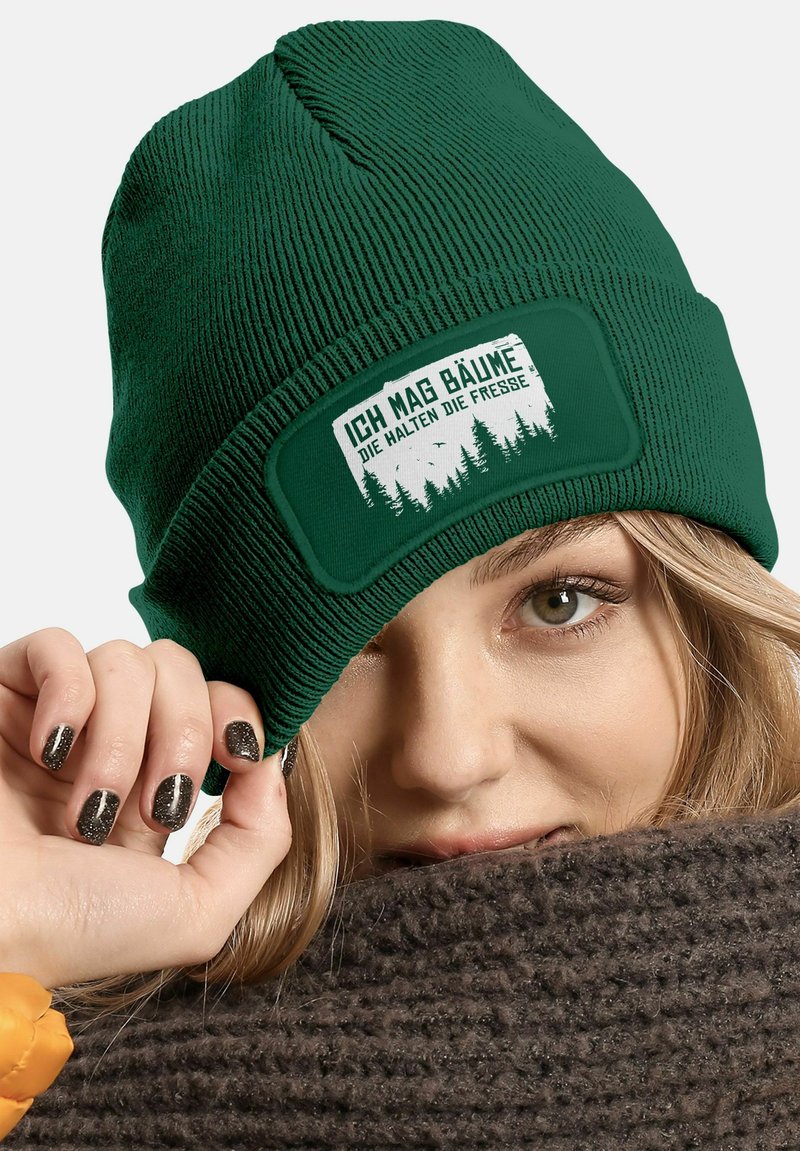 Green knit beanie with a textured finish. Features a rectangular patch with white lettering and forest graphic. Worn just above the brow.