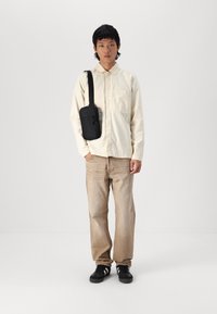 Beige button-up shirt, light brown jeans, and black sneakers. A small black crossbody bag with a zip is worn on the left shoulder.