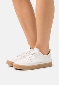 Baskets en cuir blanc avec un bout arrondi, semelle en caoutchouc marron terre et lacets blancs. Texture lisse avec des détails de couture minimes.