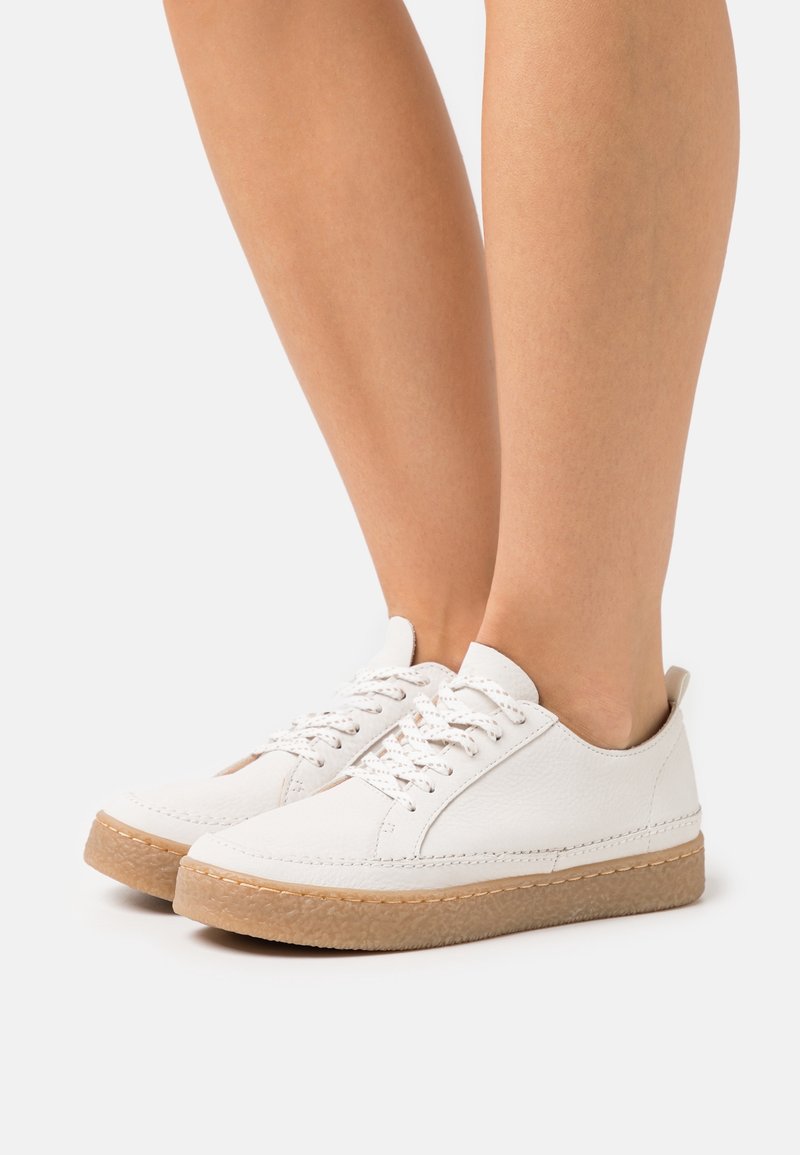 Baskets en cuir blanc avec un bout arrondi, semelle en caoutchouc marron terre et lacets blancs. Texture lisse avec des détails de couture minimes.