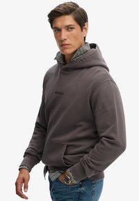 Sudadera gris con un bolsillo tipo canguro, que presenta un logotipo sutil. Lleva encima una camisa de cuadros con cuello, combinada con jeans de mezclilla azules.