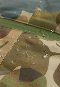 Vandafvisende stof med camouflage-mønster i jordfarver. Har dråber på overfladen og en grøn lynlås langs sømmen.