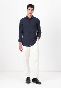 Marineblaues Langarmhemd mit Knopfverschluss, kombiniert mit weißen Hosen und schwarzen Schuhen. Der Stoff wirkt glatt; das Hemd hat eine taillierte Silhouette.