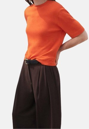 Persoon draagt een oranje top met korte mouwen, ingestopt in een donkerbruine wijde broek met een hoge taille en een zwarte riem.