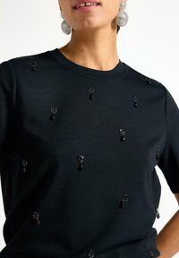 Sort kortærmet top med teksturerede udsmykninger med sorte perler og dråpe accenter, sat mod en glat stofoverflade.