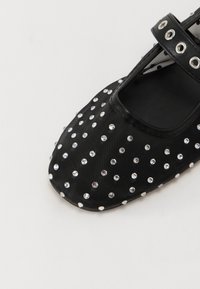 Chaussure Mary Jane noire avec une surface en tissu lisse ornée de strass dispersés, à bout rond et avec une bride à la cheville.