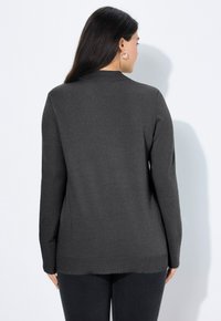 Frau mit langen dunklen Haaren, die einen dunkelgrauen Langarmpullover und schwarze Hosen trägt, mit dem Rücken zu einer weißen Wand.