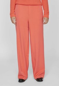 Žena v korálových kalhotách s širokými nohavicemi, odpovídajícím texturovaným svetrem a černými botami stojí na jednobarevném šedém pozadí.