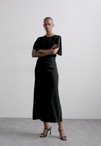 Rochie neagră, până la podea, cu mâneci scurte și drapate; textură netedă și siluetă ajustată. Asortată cu sandale negre cu fâșii.
