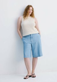 Femme aux cheveux roux bouclés portant un haut en maille crème sans manches, un short bleu clair jusqu'aux genoux et des tongs noires, debout contre un mur blanc.