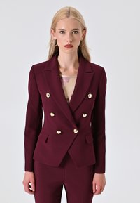 Blazer bordeaux doppiopetto con bottoni dorati, due tasche frontali e un design strutturato. Indossato sopra un top chiaro a fantasia.