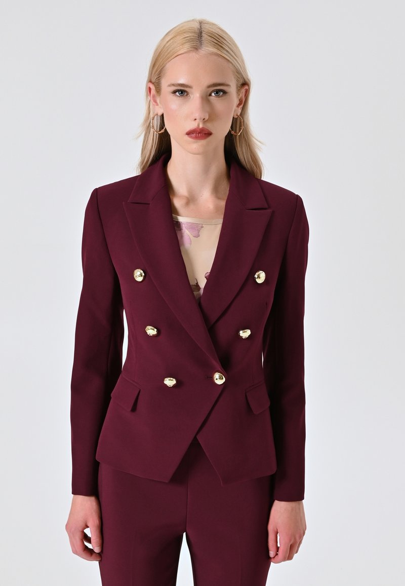 Blazer bordeaux doppiopetto con bottoni dorati, due tasche frontali e un design strutturato. Indossato sopra un top chiaro a fantasia.