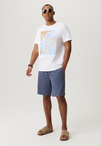 Biała bawełniana koszulka z niebieską i beżową grafiką palm; granatowe szorty; beżowe klapki; okulary przeciwsłoneczne aviator; pozycja stojąca.