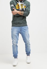 Hombre con sudadera verde de Superdry, pantalones deportivos de mezclilla azul claro, zapatillas blancas, con los brazos cruzados mostrando un tatuaje en el antebrazo izquierdo.
