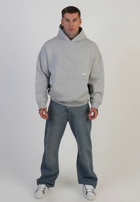 Grå bomullshoodie med framficka, kombinerad med ljusblå denimjeans. Modellen står mot en neutral bakgrund med sneakers.