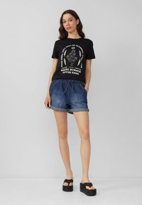 T-shirt graphique noir avec texte et design blancs, associé à un short en denim bleu avec une taille élastique et des ourlets retroussés. Sandales à plateforme noires.