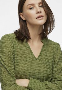 Grøn, struktureret sweater med V-hals og lange ærmer, der har et bølgende mønster. Stoffet ser let og en smule gennemsigtigt ud.