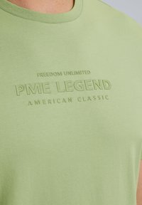 Svetle zelené bavlněné tričko s kulatým výstřihem a embosovaným textem: "NEOMEZENÁ SVOBODA PME LEGEND AMERICAN CLASSIC" vpředu.