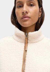 Kremowa polarowa kurtka zapinana na zamek błyskawiczny z wysokim kołnierzem, z metalowym suwakiem i skórzaną pętelką w kolorze brązowym. Miękka faktura, minimalistyczny design.