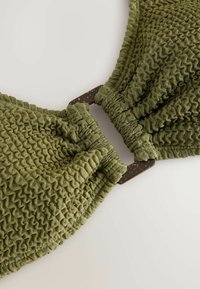Tissu texturé vert olive avec des détails froncés, comportant une fermeture rectangulaire marron foncé comme accent principal.