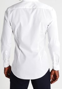Vit långärmad skjorta med slät textur, rundad fåll och knäppta manschetter. Bakifrån syns sömmarna och en klassisk krage.