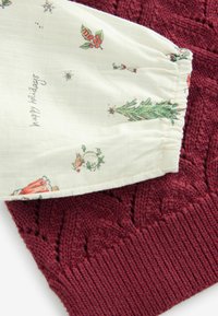 Manchon en tissu blanc avec des illustrations sur le thème des fêtes et le texte "joyeuses fêtes" sur un fond texturé en tissu tricoté rouge foncé.