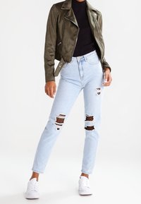 Chaqueta de moto de ante sintético verde oliva, camiseta negra, jeans claros desgastados con malla en dessous y zapatillas blancas.