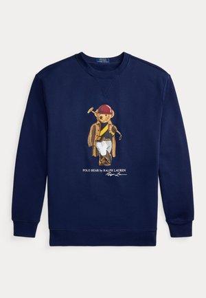 Mand med langt krøllet hår iført marineblå sweatshirt med teddybjørnegrafik, hvide bukser og hvide sneakers, stående foran en neutral baggrund.