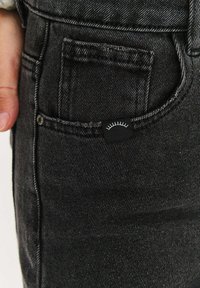 Černé džínové kalhoty s přední kapsou, na které je malá černá etiketa s minimalistickým bílým motivem. Textura působí odolně.