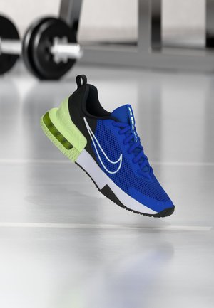 Niebiesko-czarny but sportowy z neonowo zielonym, powietrznym amortyzowanym obcasem, białą środkową podeszwą oraz dużym białym logo Nike, unoszący się w siłowni.