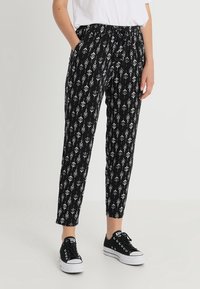 Schwarz gemusterte Hose mit einem weißen geometrischen Design, elastischem Bund und seitlichen Taschen, kombiniert mit schwarzen Sneakers.