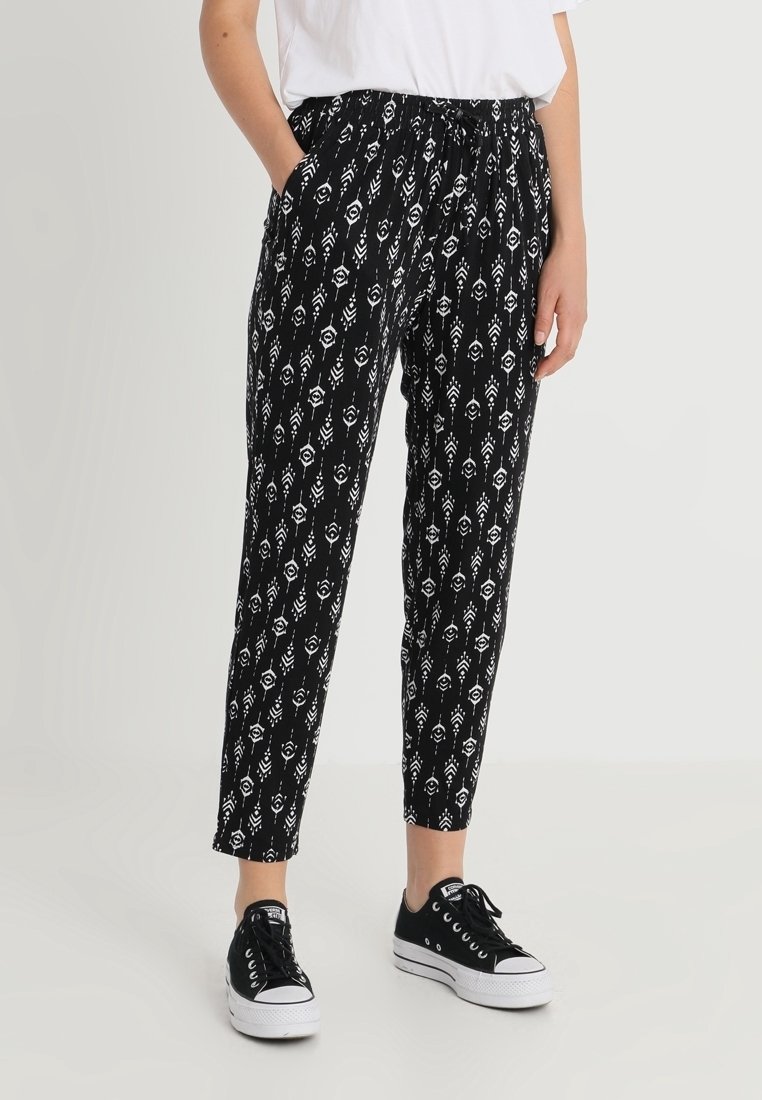 Schwarz gemusterte Hose mit einem weißen geometrischen Design, elastischem Bund und seitlichen Taschen, kombiniert mit schwarzen Sneakers.