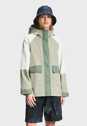 Ung person iført en grøn og beige hættetrøje, navy fiskerhat og sorte cargoshorts, stående mod en ensartet lys baggrund.