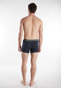 Navy blue boxers with a patterned fabric and a black waistband featuring logo text. The model stands barefoot, showing the back view.