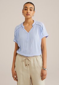 Ljusblå kortärmad blus med V-ringning, prydd med dekorativ sömnad vid axlarna, tillsammans med beige byxor med dragsko.